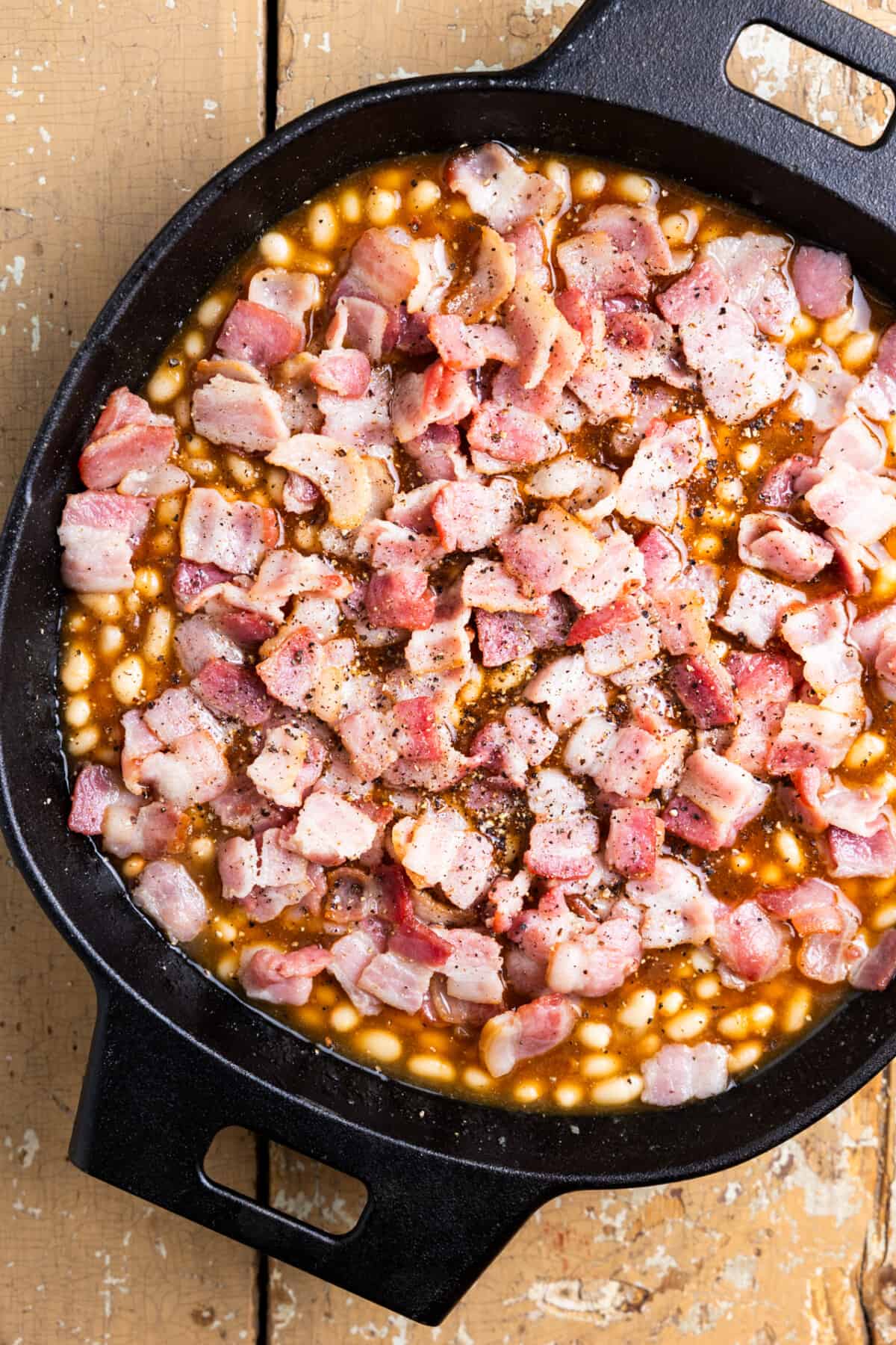 Baked beans in a skillet topped with bacon before cooking.