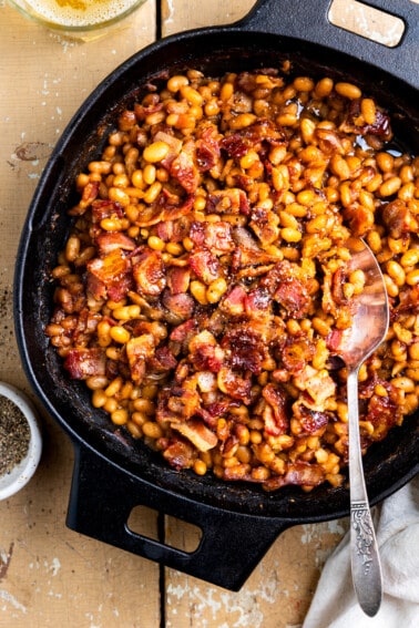 Cast iron skillet filled with baked beans topped with crispy bacon and coarse ground black pepper.