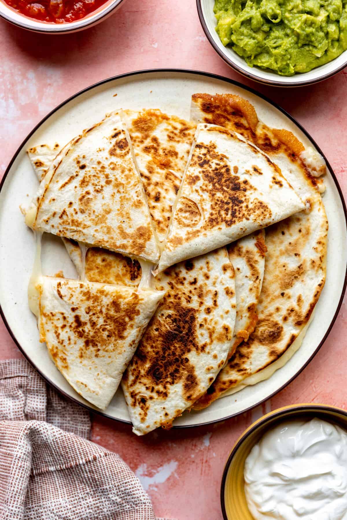 Plate filled with toasted cheese quesadillas and bowls of sour cream, guacamole and salsa on the side. 