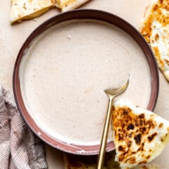 Red bowl with a cream sauce, a spoon, and a wedge of quesadilla.