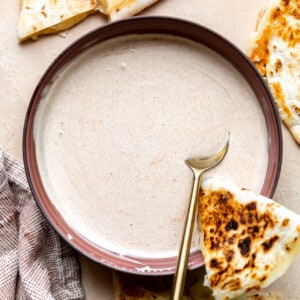 Red bowl with a cream sauce, a spoon, and a wedge of quesadilla.