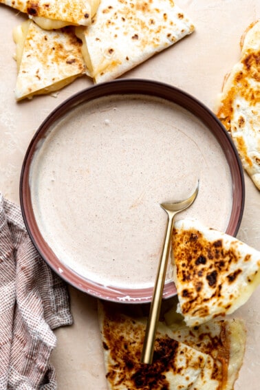 Red bowl with a cream sauce, a spoon, and a wedge of quesadilla.