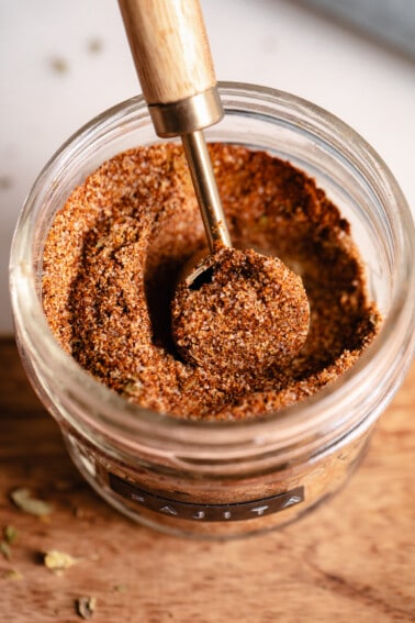 Jar filled with red spice mix with a wooden handled teaspoon scooping it up.