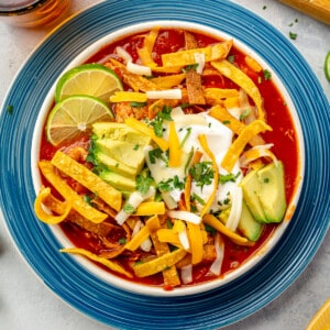 Bowl filled with chicken tortilla soup, topped with crispy tortilla strips, avocado, sour cream and slices of lime.
