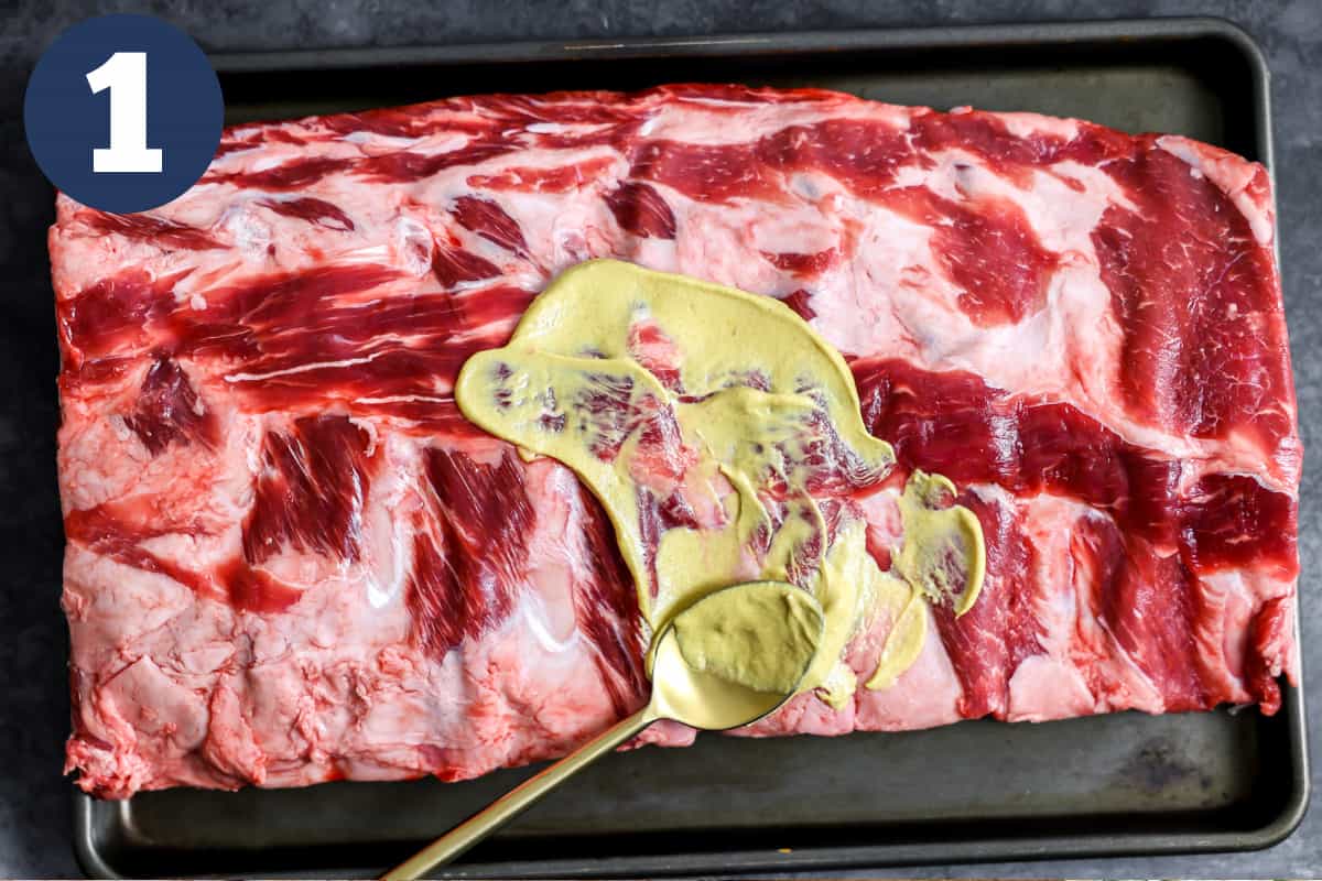 Spreading mustard on beef ribs on a baking tray.
