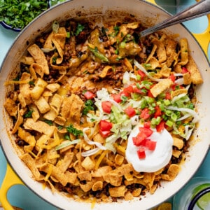 Skillet with walking taco casserole in it, topped with shredded lettuce, sour cream, and diced tomato.