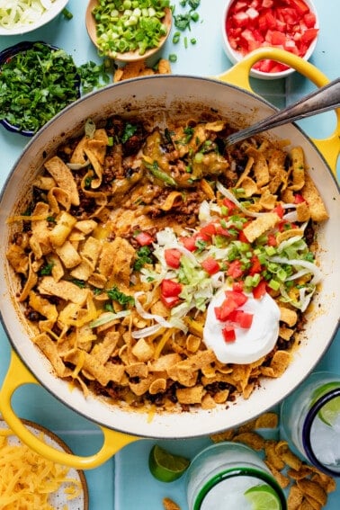 Skillet with walking taco casserole in it, topped with shredded lettuce, sour cream, and diced tomato.