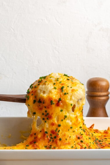 Wooden spoon scooping up mashed potatoes topped with melty cheese and diced chives.