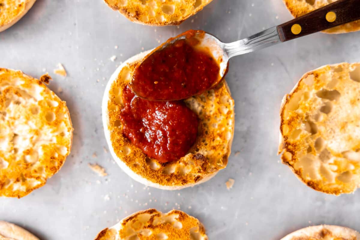 Spoon spreading pizza sauce on toasted English muffin.