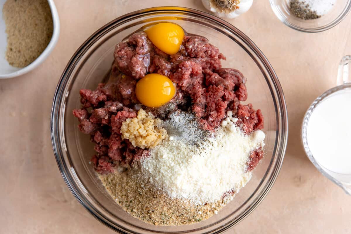 Ground beef in a bowl with bread crumbs, parmesan, eggs, garlic, and seasonings. 
