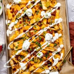 Golden, crispy French fries loaded with melted cheese, bacon, green onions, and a drizzle of sour cream on a baking tray.